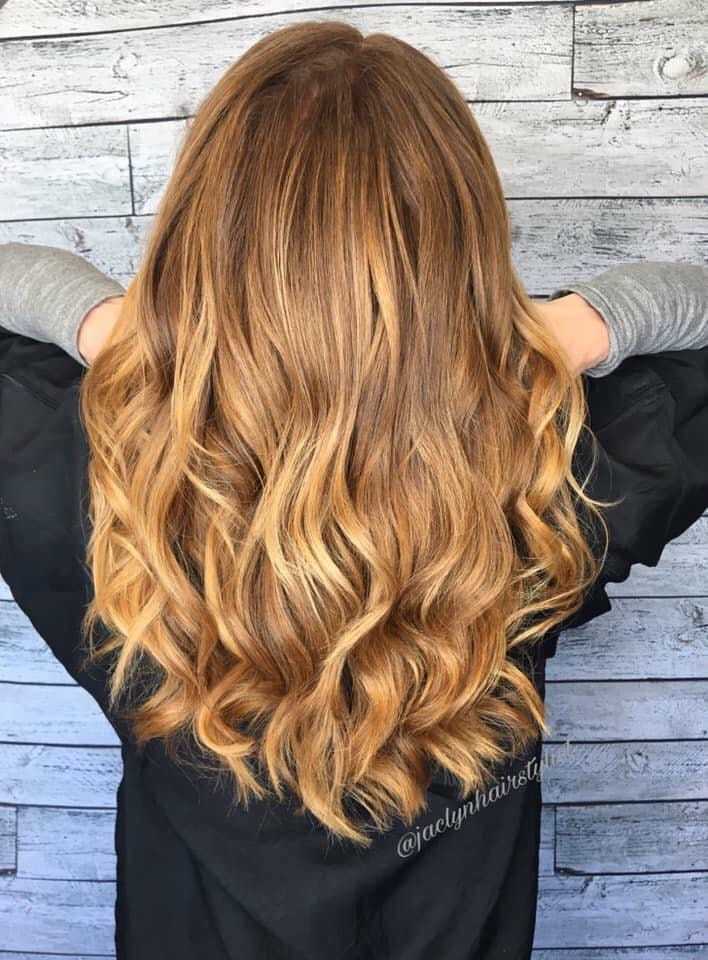 Woman with wavy, golden-brown hair facing away, hands touching head, wooden background.